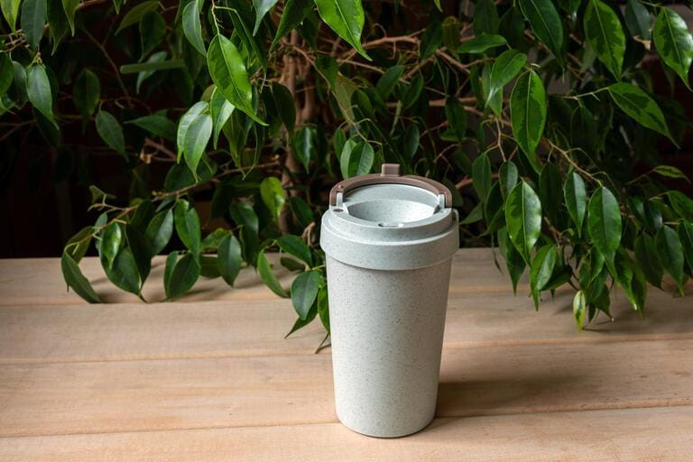 Plastic reusable thermostatic cups for coffee, tea and other hot and cold drinks on a wooden desk against a background of green foliage. 