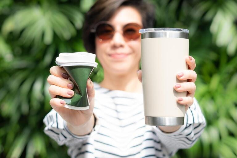A stylish beautiful woman holds a takeaway coffee cup in both hands, one is a single use paper cup with a plastic lid, the other is a reusable stainless cup. A stylish beautiful woman holds a takeaway coffee cup in both hands, one is a single use paper cup with a plastic lid, the other is a reusable stainless cup.