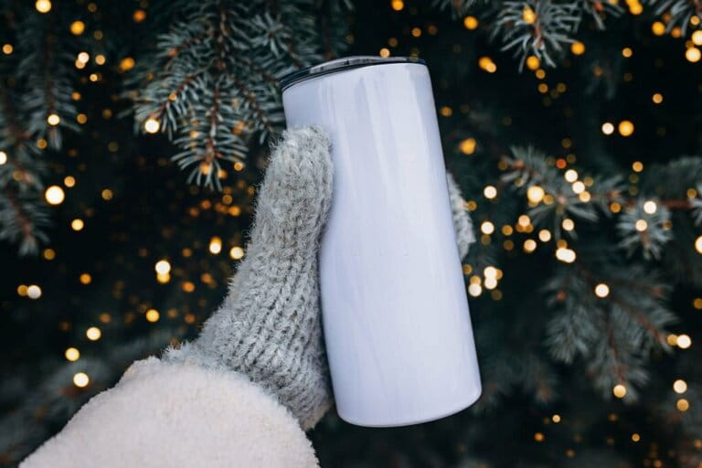 Empty white steel cup in hand on Christmas tree background. White steel tumbler mockup.