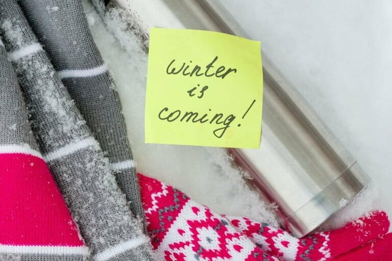 Winter is coming, text on sticker. Winter background - in the snow a scarf, gloves, thermos.
