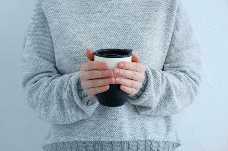 Thermal cup with hot coffee in the hands of white woman in a sweater.